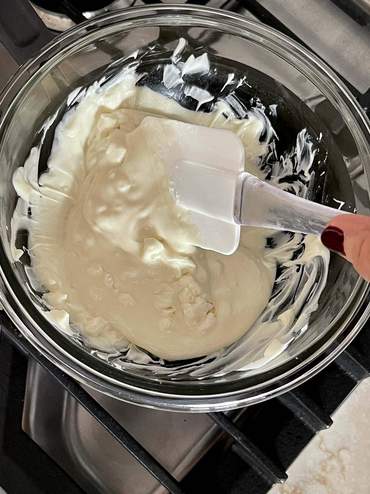 A white rubber spatula stirring melted white chocolate in a glass bowl on a stovetop.