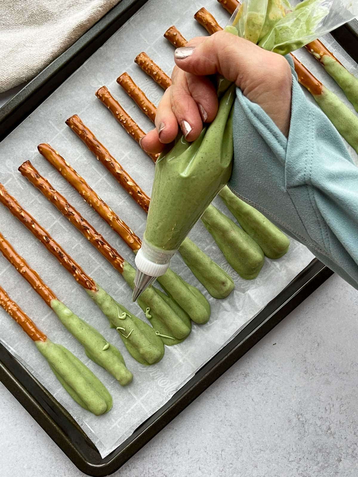 A woman holding a piping bag filled with matcha chocolate piping it over a row of pretzel rods on top of parchment paper on a black baking tray.