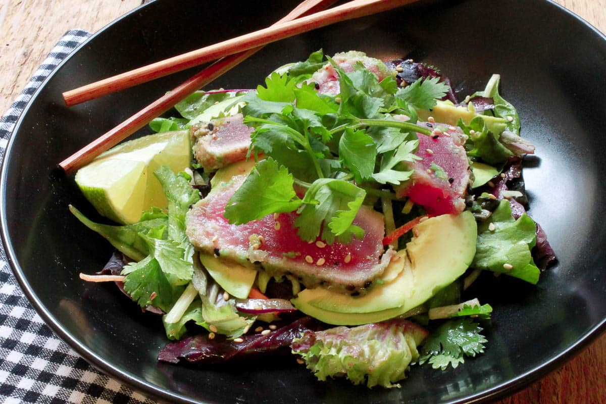 Seared ahi tuna on a bed of greens in a black bowl with wooden chopsticks on the side.