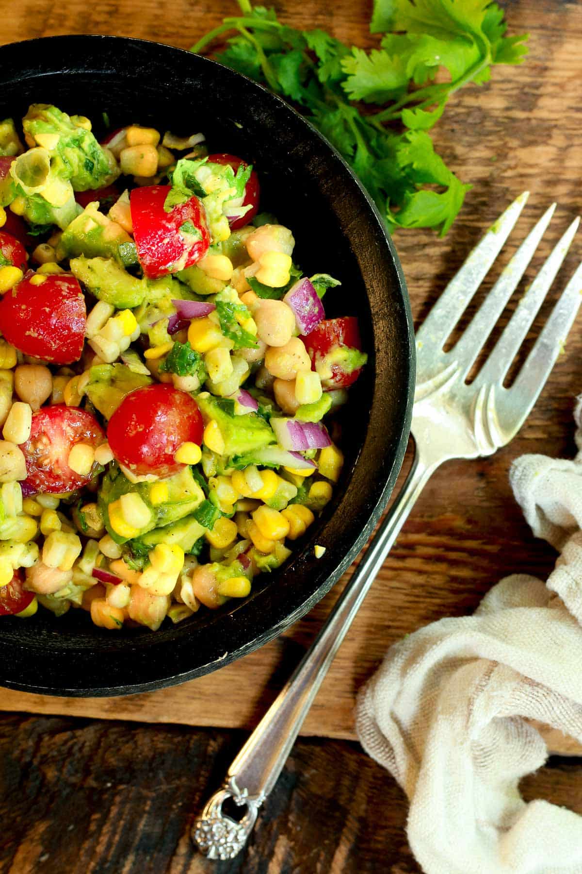 A black bowl with sweet corn avocado salad with cherry tomatoes and fresh cilantro on a wooden board with a silver form on the side.