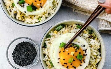 Crispy rice with ginger and scallions topped with a runny fried egg in two small bowls with a pair of brown chopsticks, a linen napkin, and black sesame seeds in a bowl on the side.