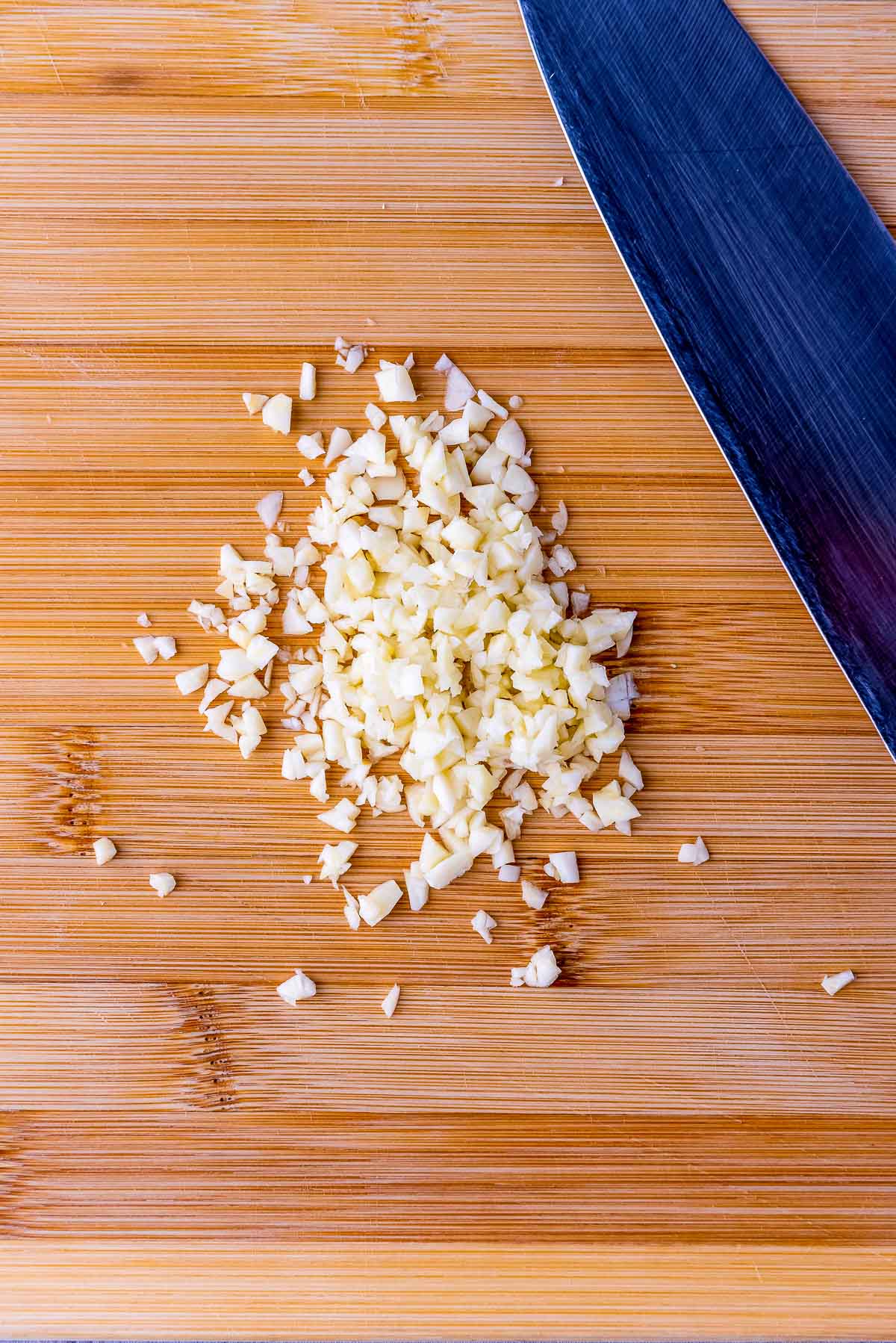 Minced garlic on a wooden cutting board with a large knife on the side.