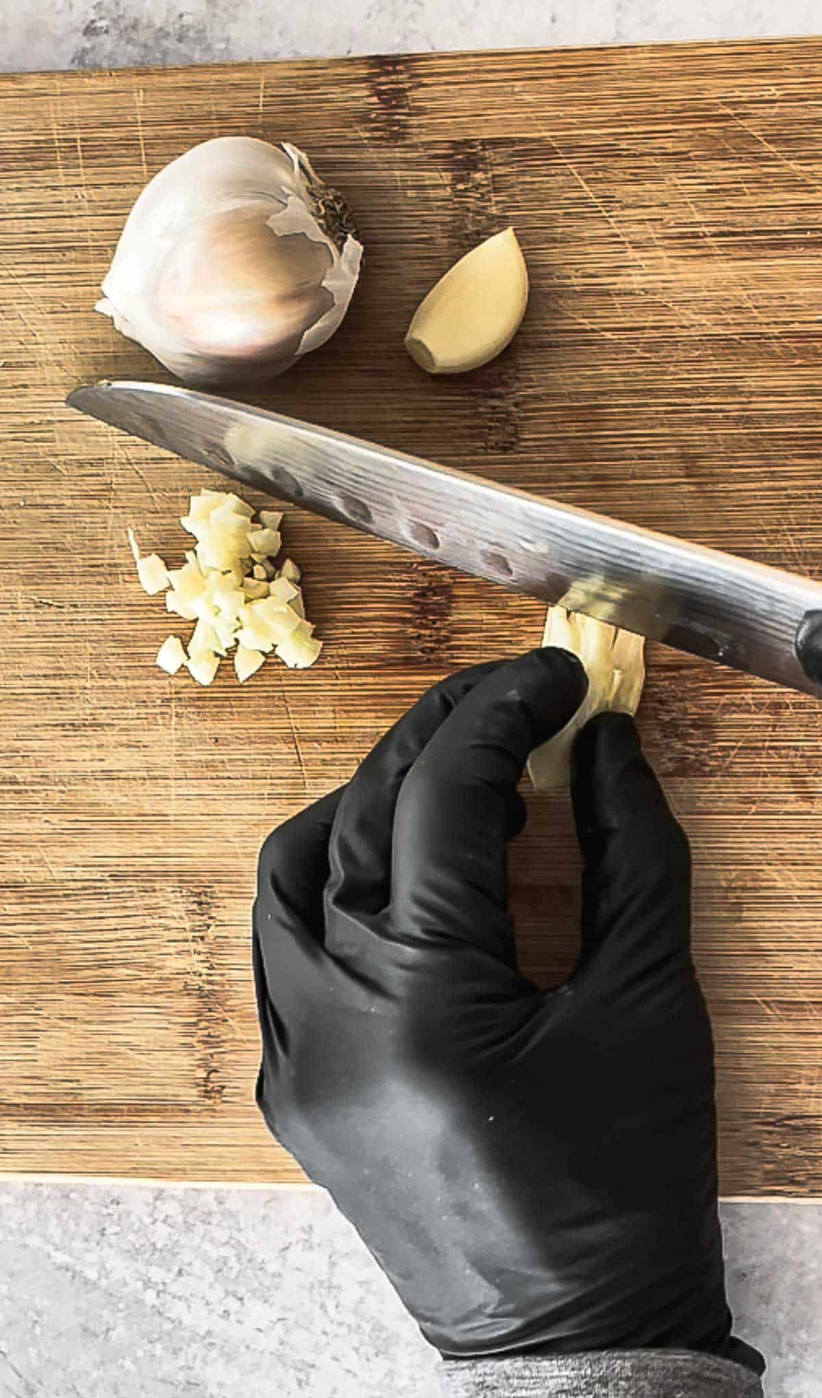 A hand with black gloves mincing fresh garlic on top of a wooden cutting board with a pile of minced garlic and a whole garlic bulb on the side.