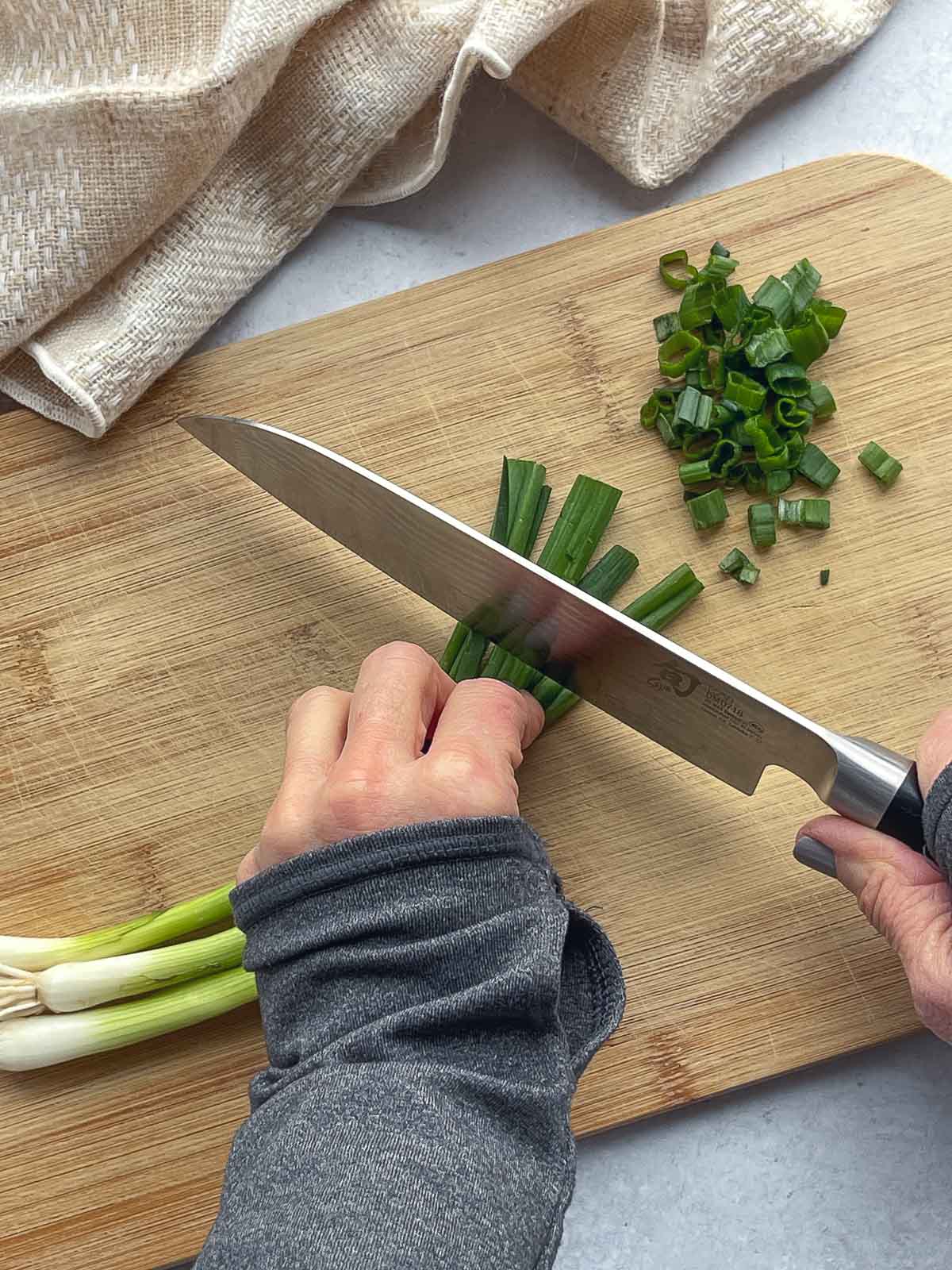 A woman's hands slicing green scallions on top of a wooden cutting boar with a linen napkin on the side.
