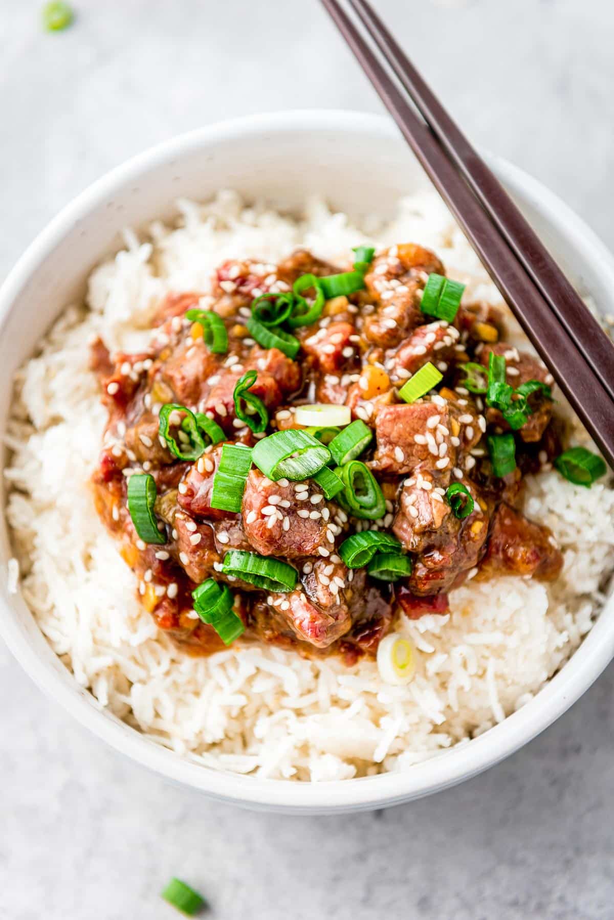 A pair of chopsticks on top of a white bowl filled with Slow Cooker Mongolian Beef on white rice on top of a gray surface.