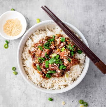A pair of chopsticks on top of a white bowl filled with Slow Cooker Mongolian Beef on white rice, with a bowl of Mongolian beef in the background on top of a gray surface.