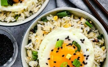 Crispy rice with ginger and scallions topped with a runny fried egg in two small bowls with a pair of brown chopsticks and black sesame seeds in a bowl on the side.