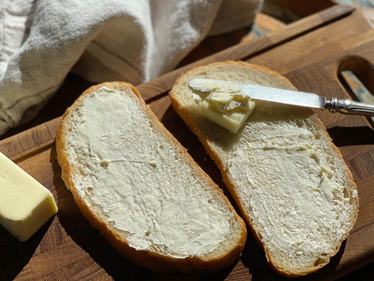 Two slices of buttered sourdough bread on top of a wooden cutting board with a butter knife and pat of butter.