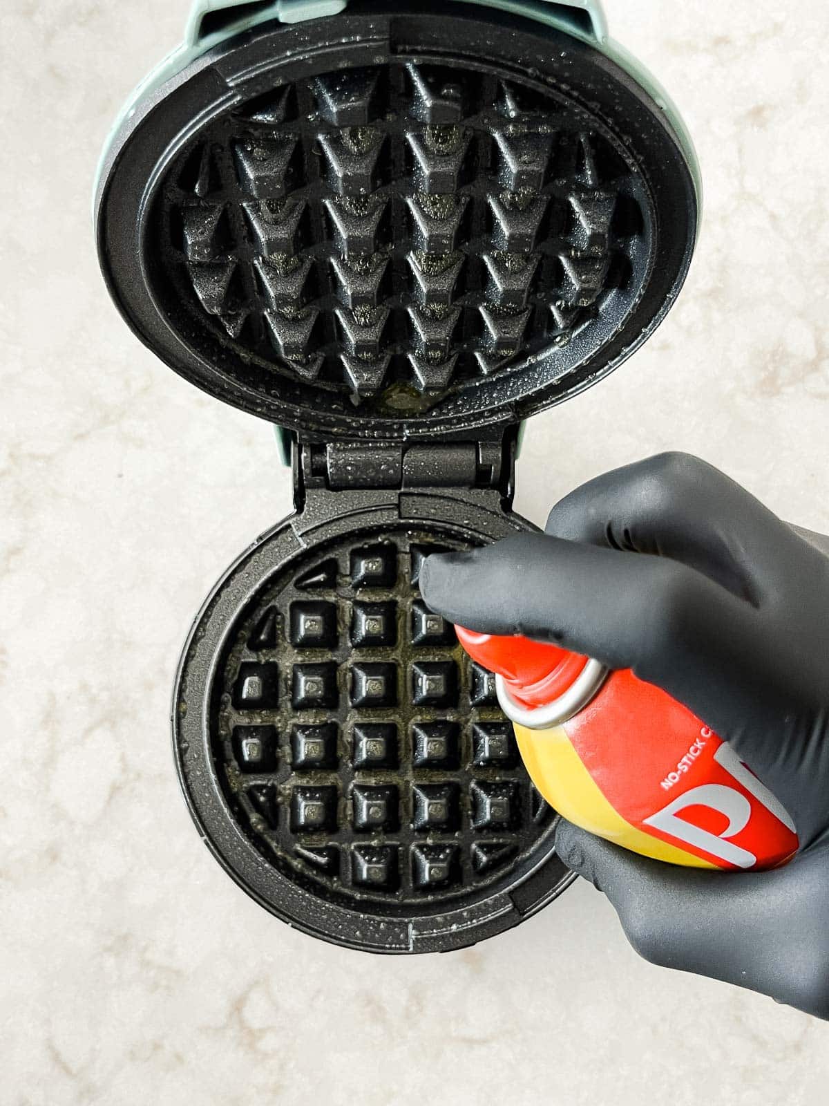 A hand spraying cooking spray on a waffle maker on top of a marble board.