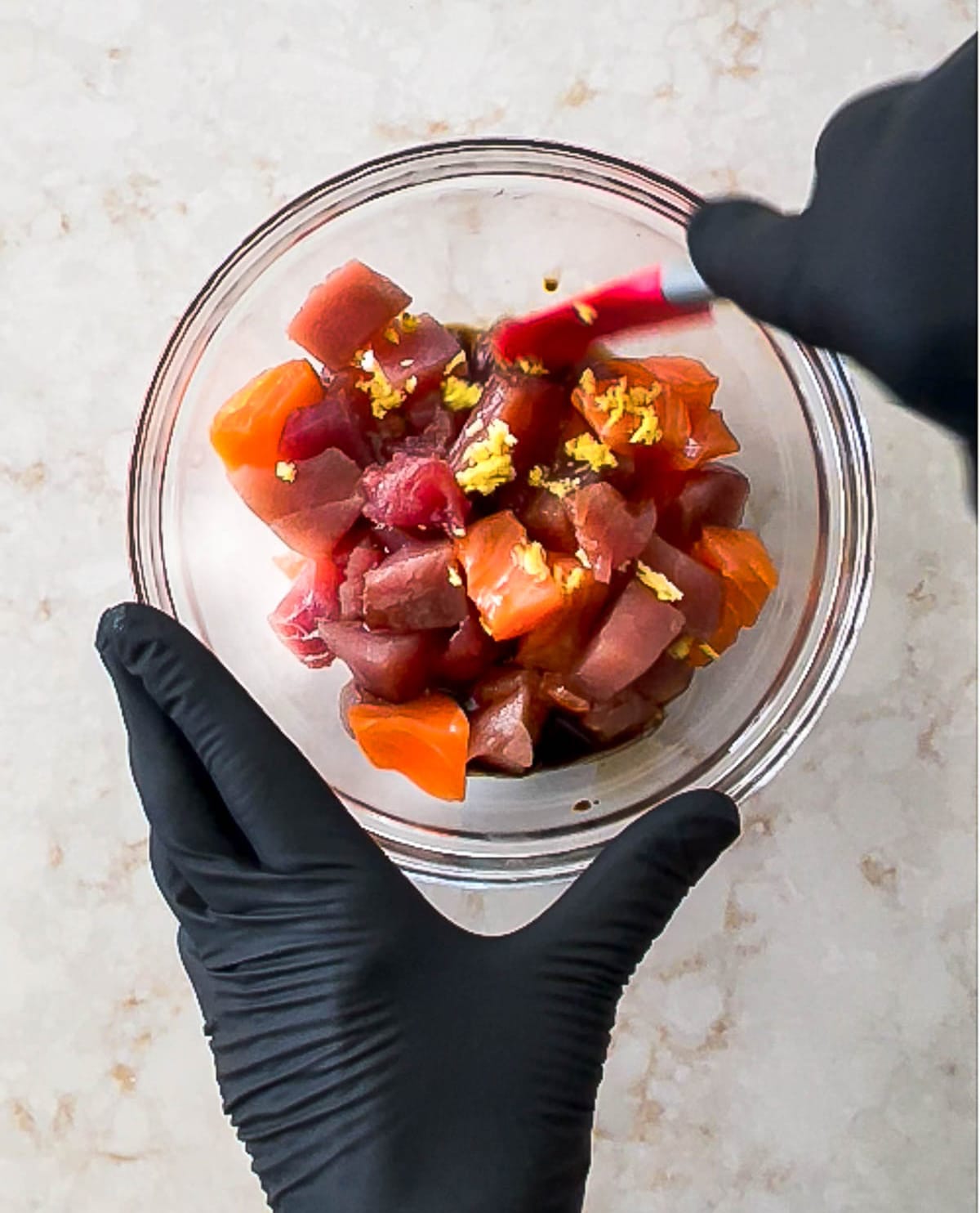Two hands mixing up cubed salmon and tuna in a glass bowl on top of a marble surface.