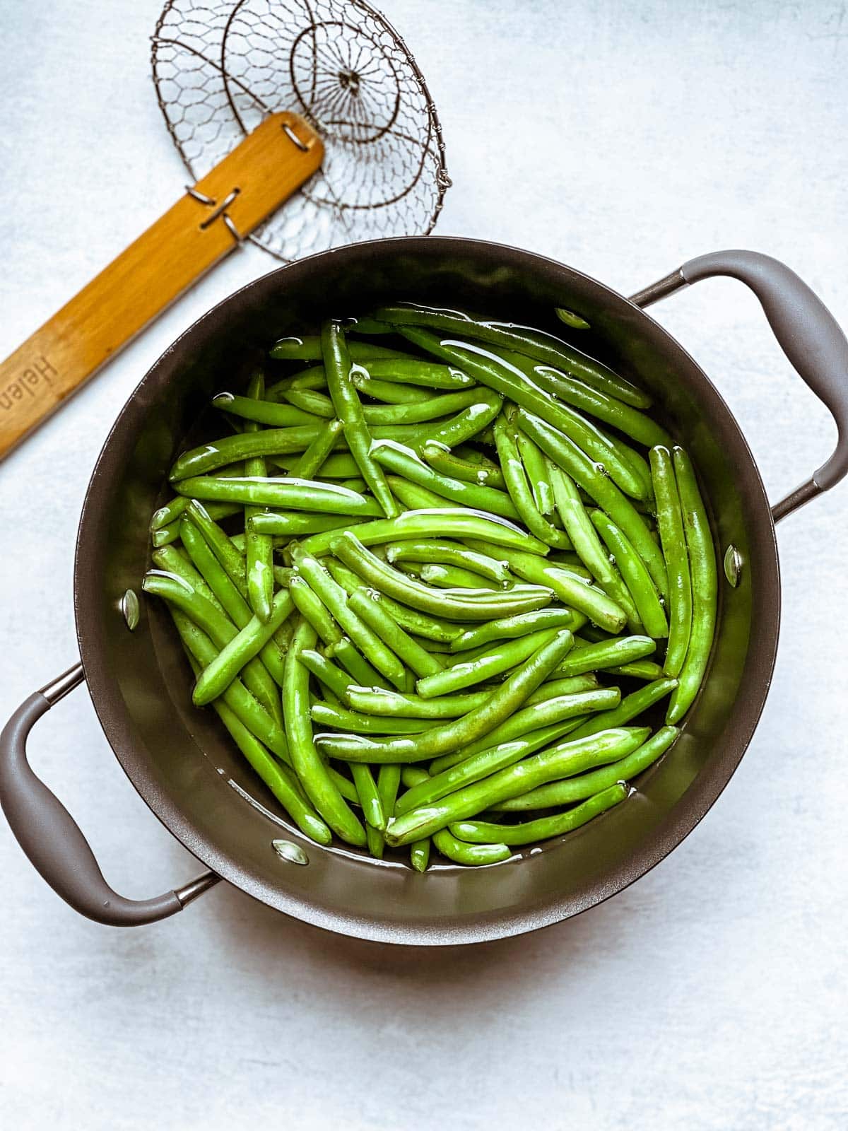 Green beans cooking in water in a pot with a spider strainer on the side.