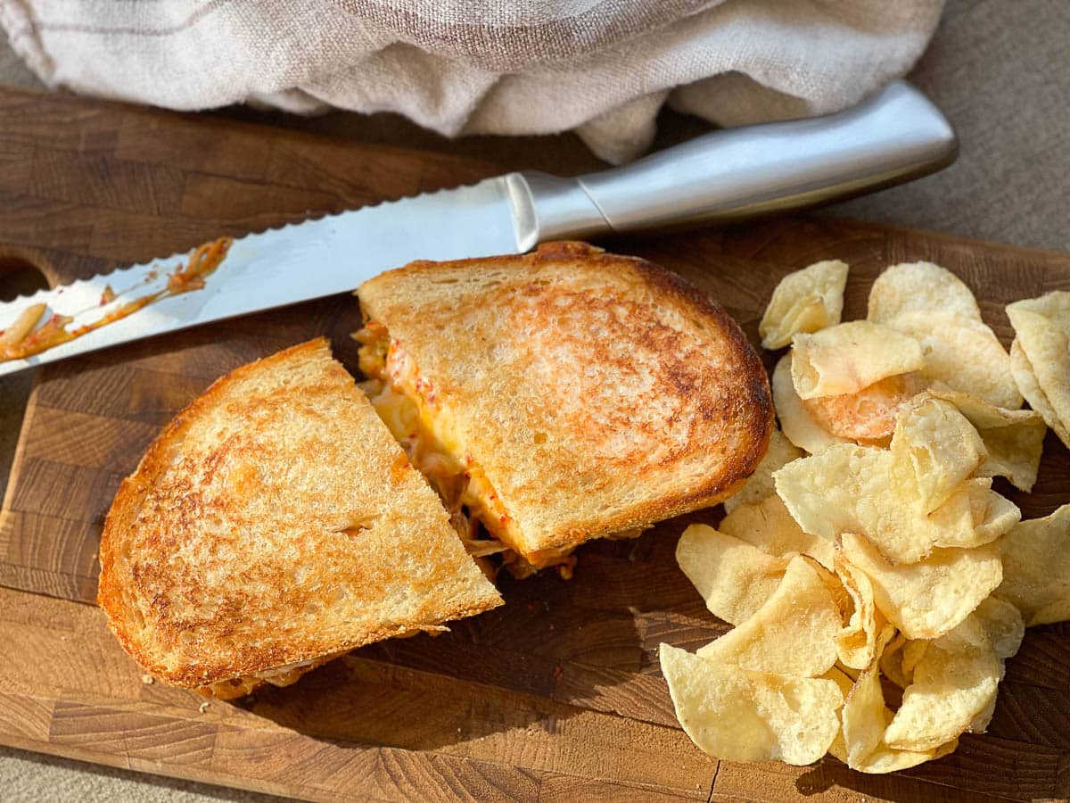 A golden grilled cheese sandwich sliced in half on a wooden board with potato chips and a cutting knife on the side.