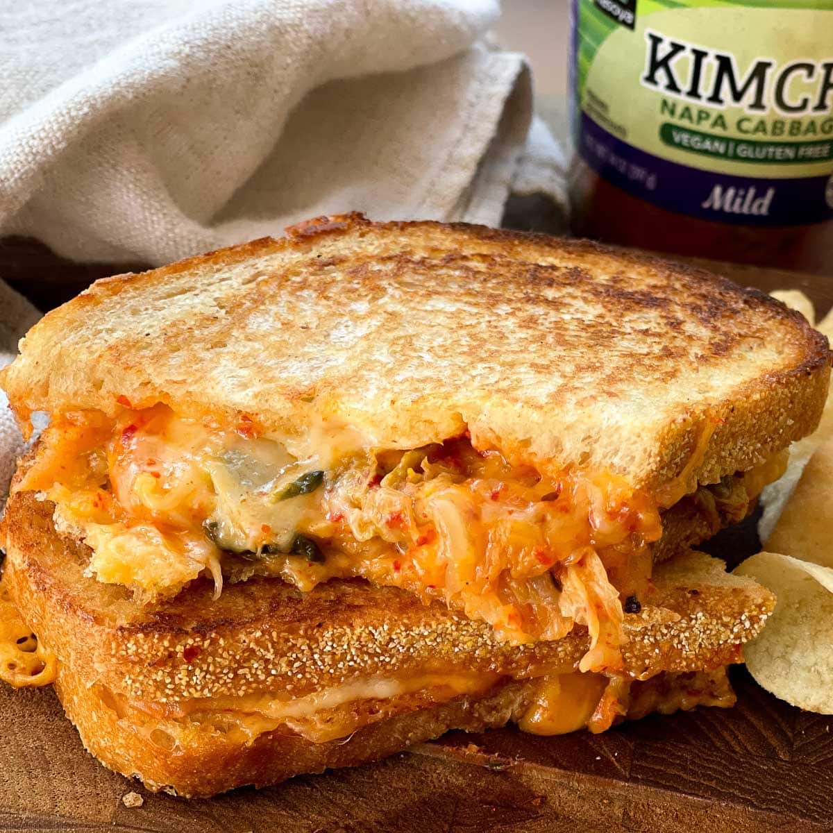 A stacked kimchi grilled cheese sandwich on a wooden board with a jar of kimchi and potato chips on the side.