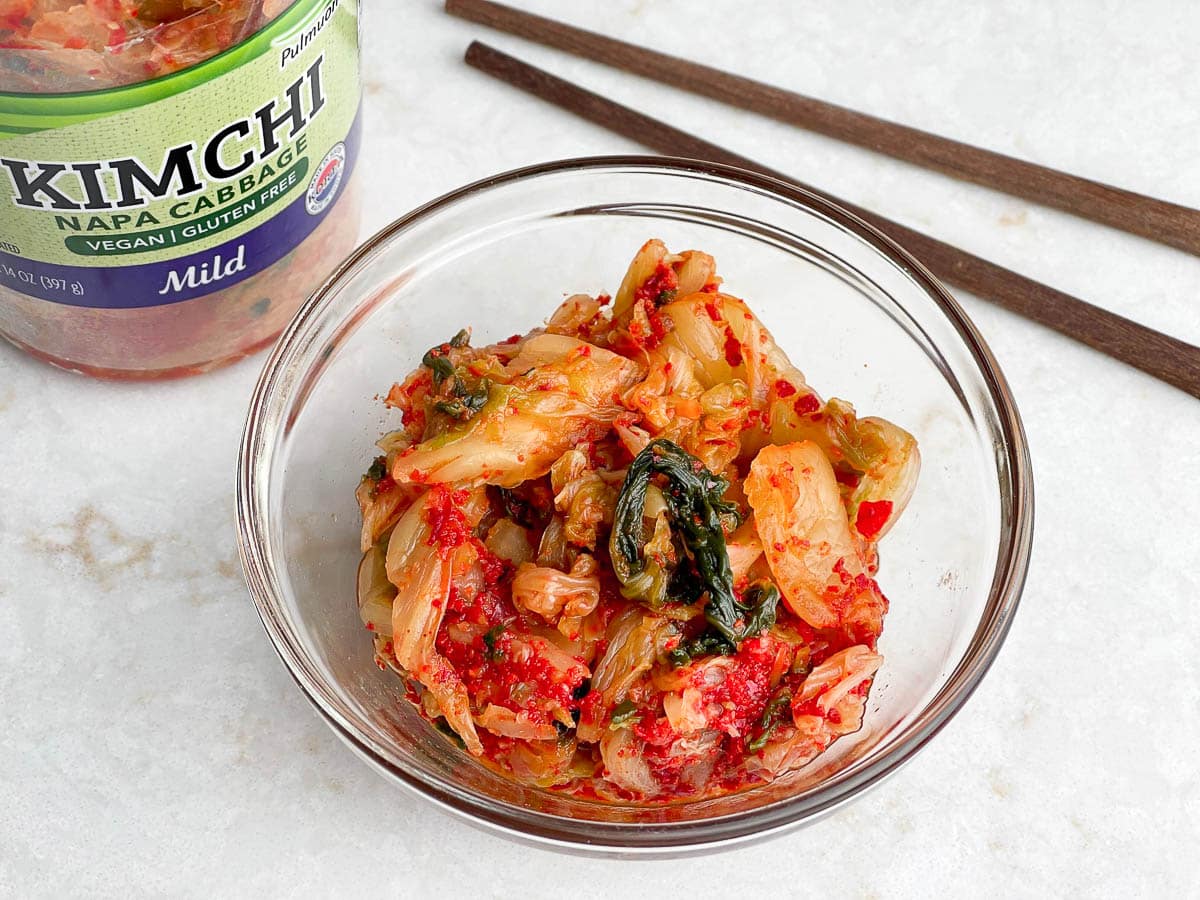Kimchi in a clear bowl with a bottle of Kimchi and a pair of chopsticks on the side.