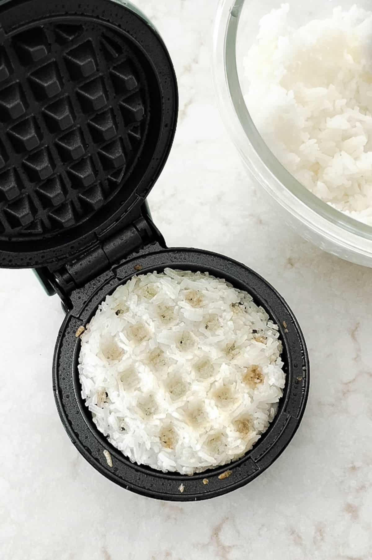 A white rice waffle in a small black waffle maker with a clear bowl of white rice on the side on top of a marble surface.