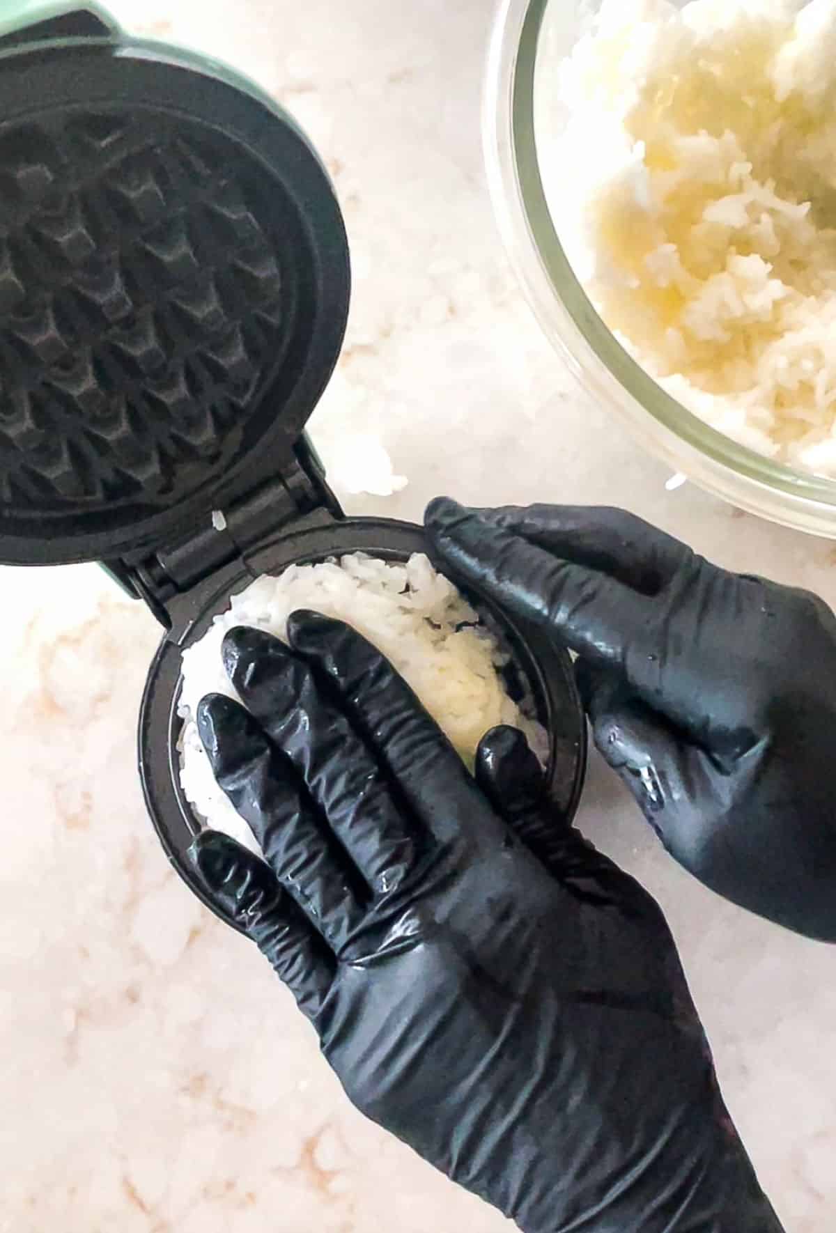 Hands pressing cooked white rice into a small black waffle maker.