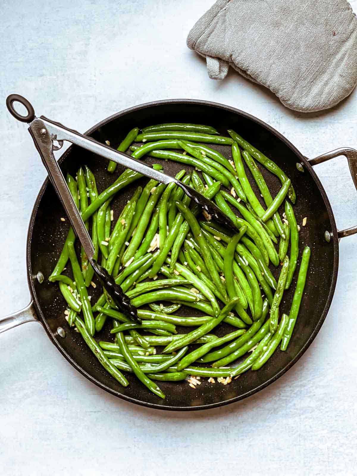 Tongs inserted in a frying pan with sauteed green beans and garlic and an oven mitt on the side.