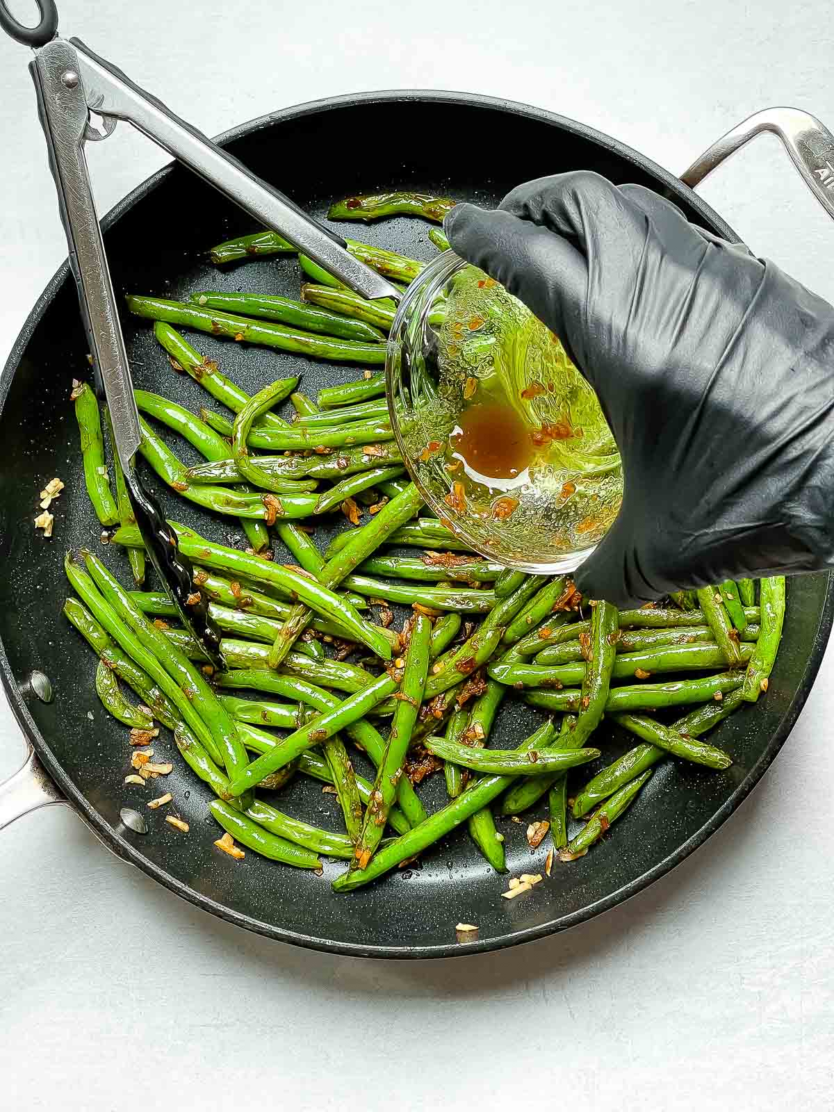 Sauce being poured on top of Chinese Garlic Green Beans sauteeing in a frying pan with tongs inserted.
