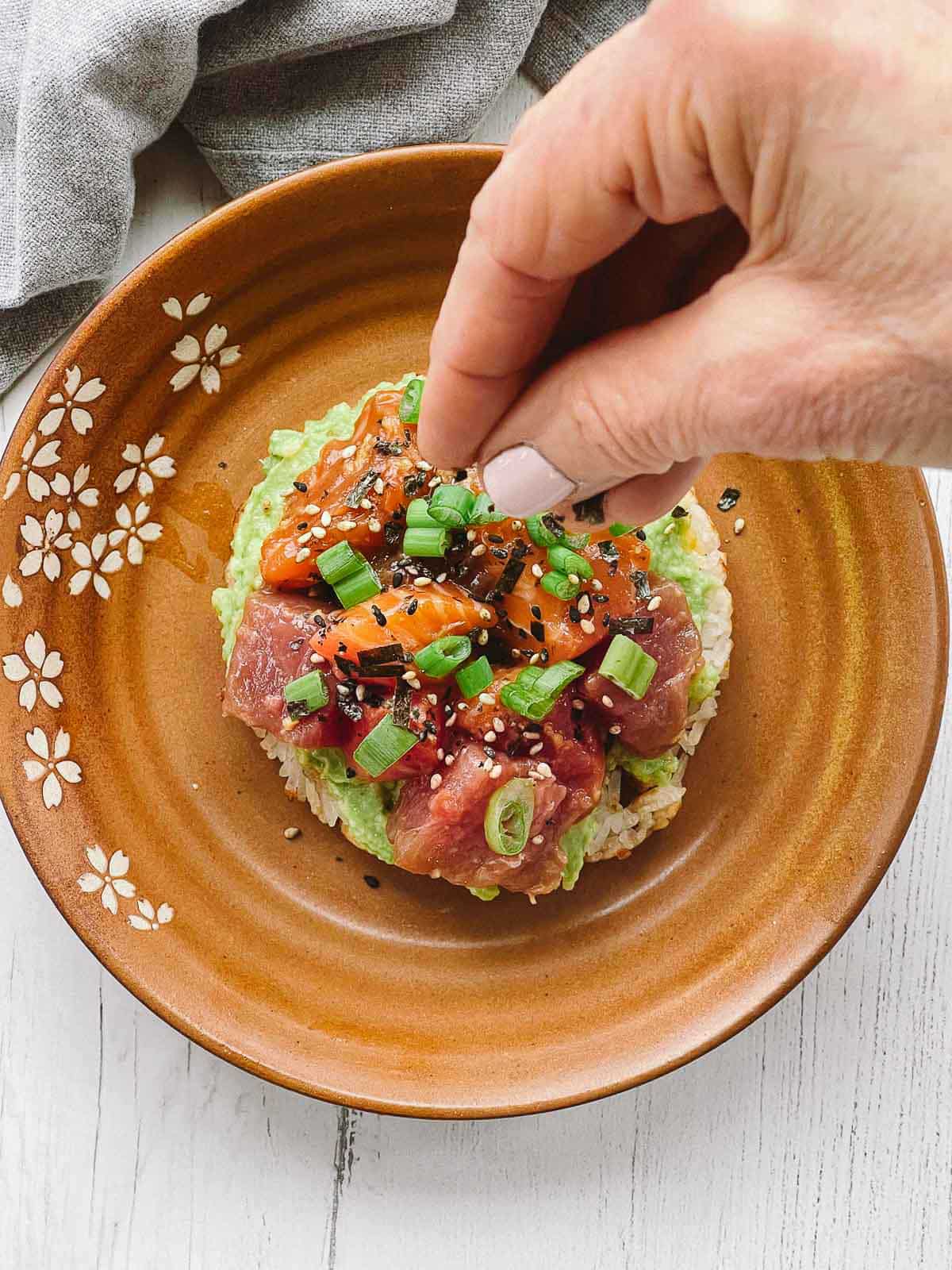 A hand sprinkling sesame seeds on top of a sushi waffle on a brown plate.