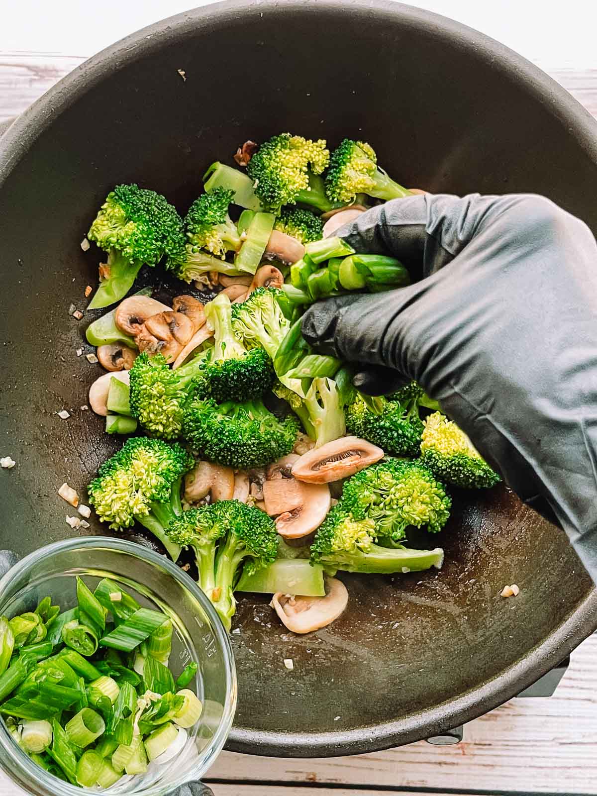 Sliced green onions in a small glass bowl being added to a wok with stir-fried broccoli florets and sliced mushrooms.