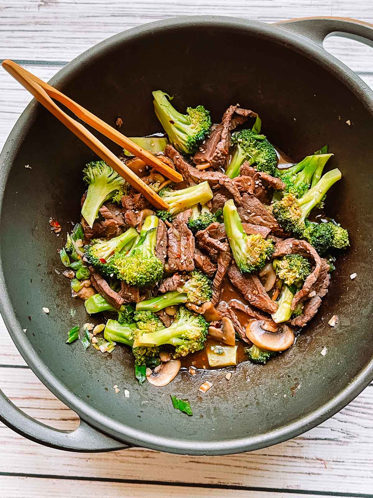 Beef and broccoli stir fry in a large wok with a pair of wooden tongs.