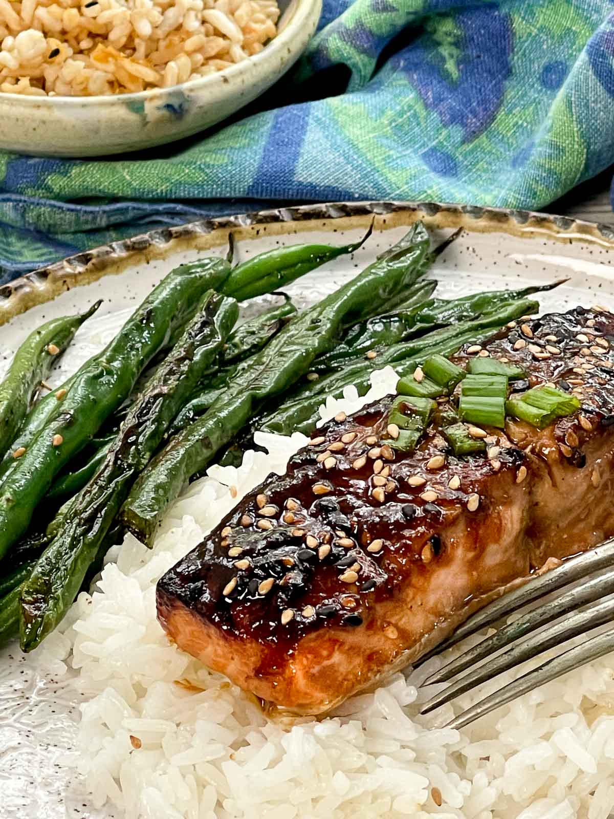 A filet of broiled miso glazed salmon on top of white rice on a plate with a fork and blistered green beans and a small bowl of brown rice and a blue napkin on the side.