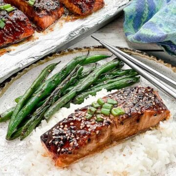 A filet of broiled miso glazed salmon on top of white rice on a plate with silver chopsticks and blistered green beans and a baking tray with broiled miso glazed salmon filets and a blue napkin on the side.