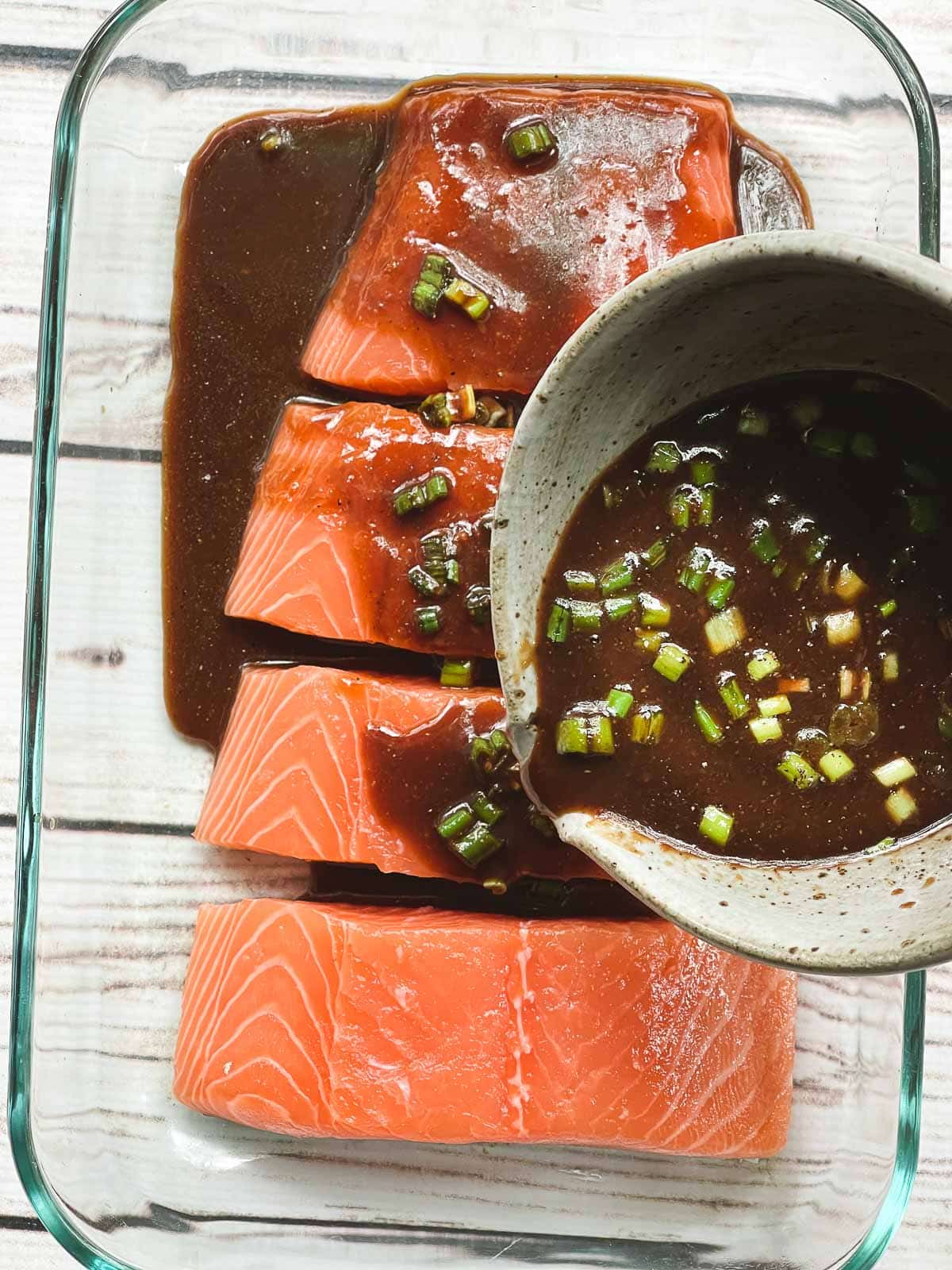 Pouring a miso marinade in a gray bowl on top of four raw salmon filets in a glass pyrex dish, on top of a white board.