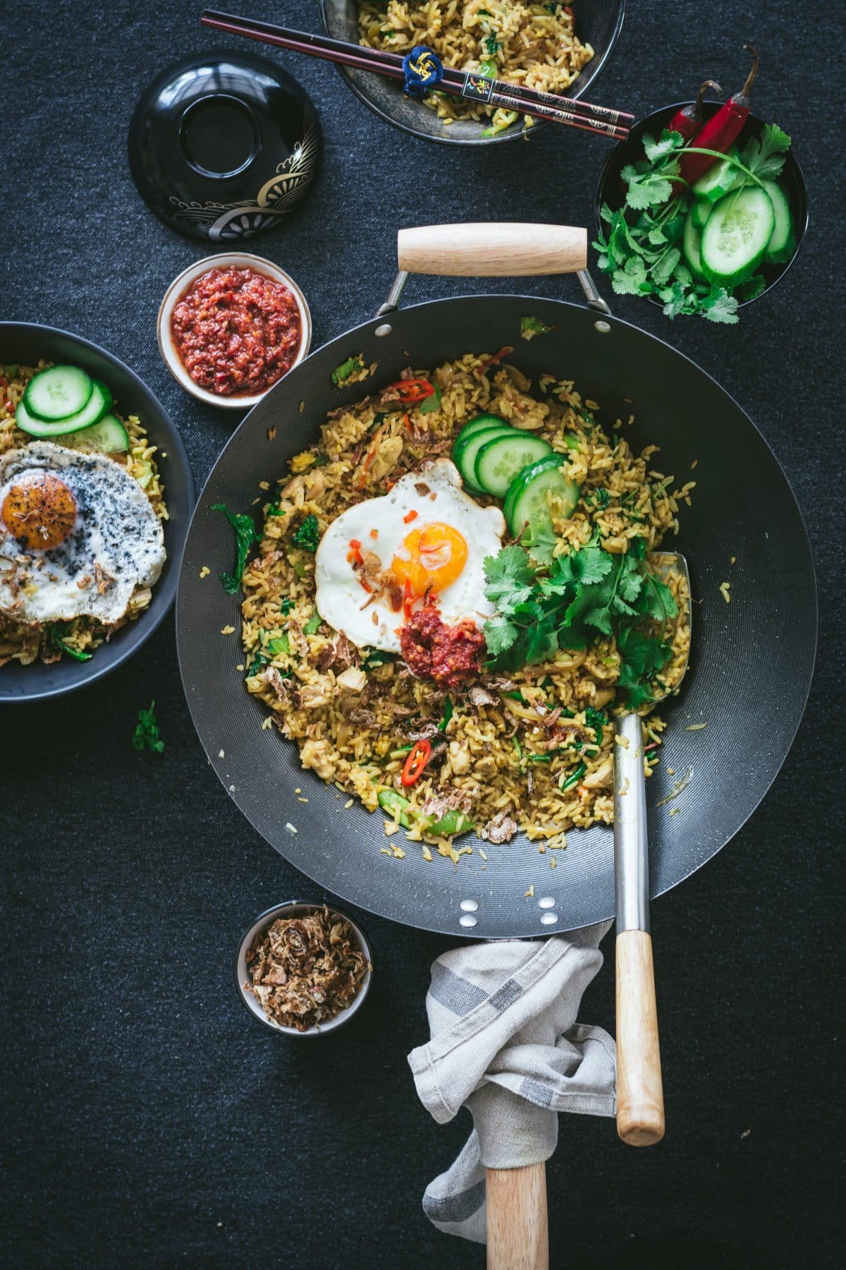 Asian chicken fried rice in a large wok with a fried egg, sliced cucumbers, and cilantro leaves on top and other condiments in small bowls on the side on top of a black surface.