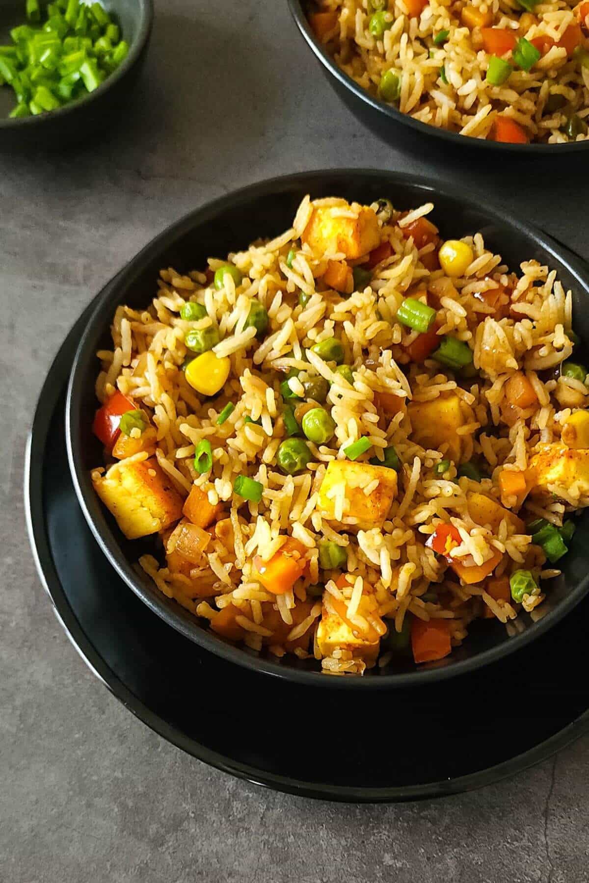 Paneer fried rice in a round black bowl with another bowl of fried rice and a small bowl of chopped green onions on the side on top of a dark gray surface.