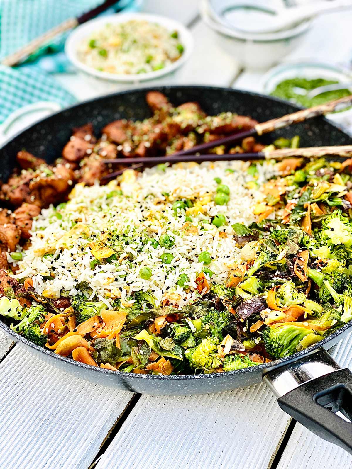 Teriyaki chicken and egg fried rice with vegetables in a large skillet topped with chopsticks on top of a white wooden board with topping in small bowls on the side.