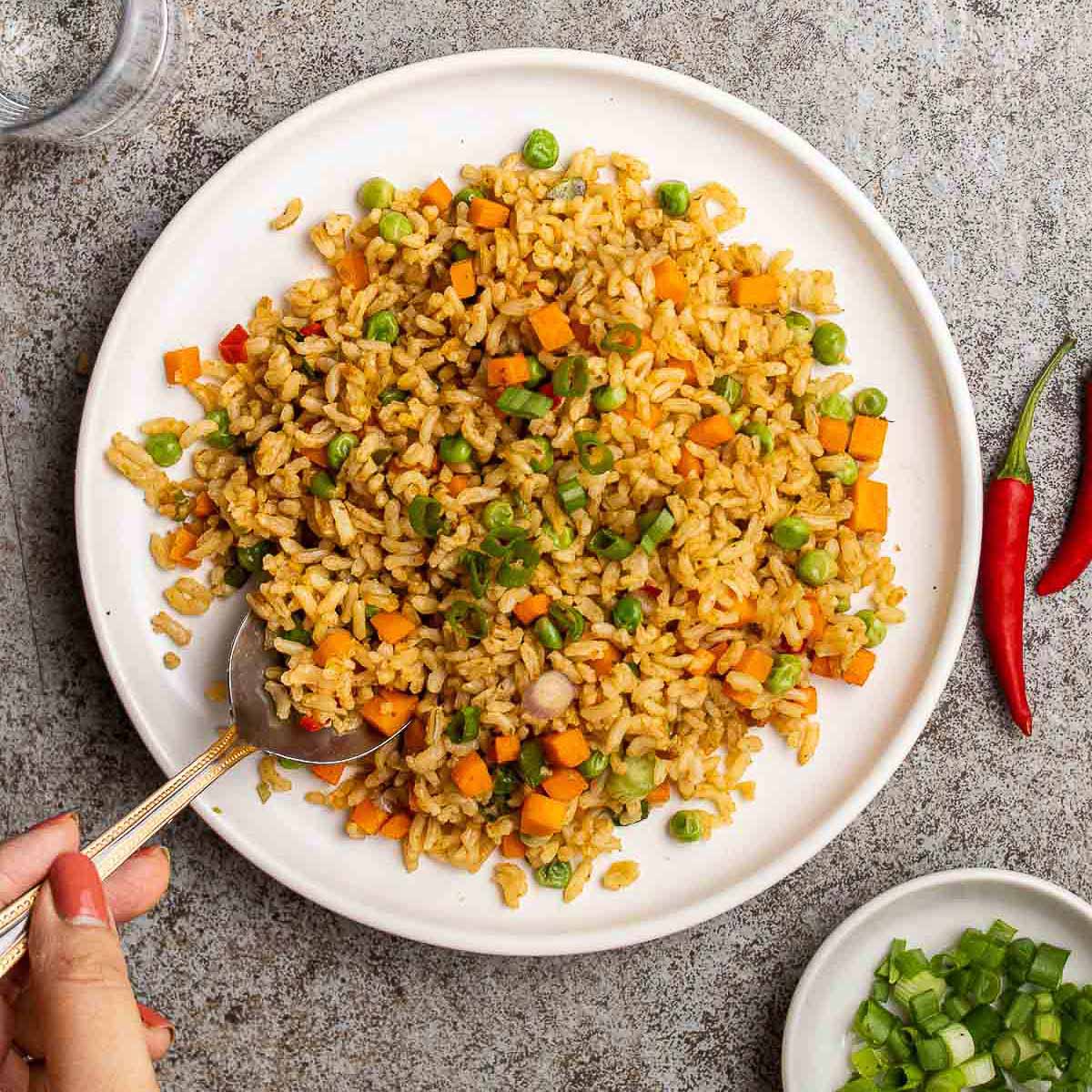 Tom Yum Fried Rice on a round white plate with a woman's hand with a spoon with red chili peppers and a small bowl of chopped green onions, on top of a gray surface.