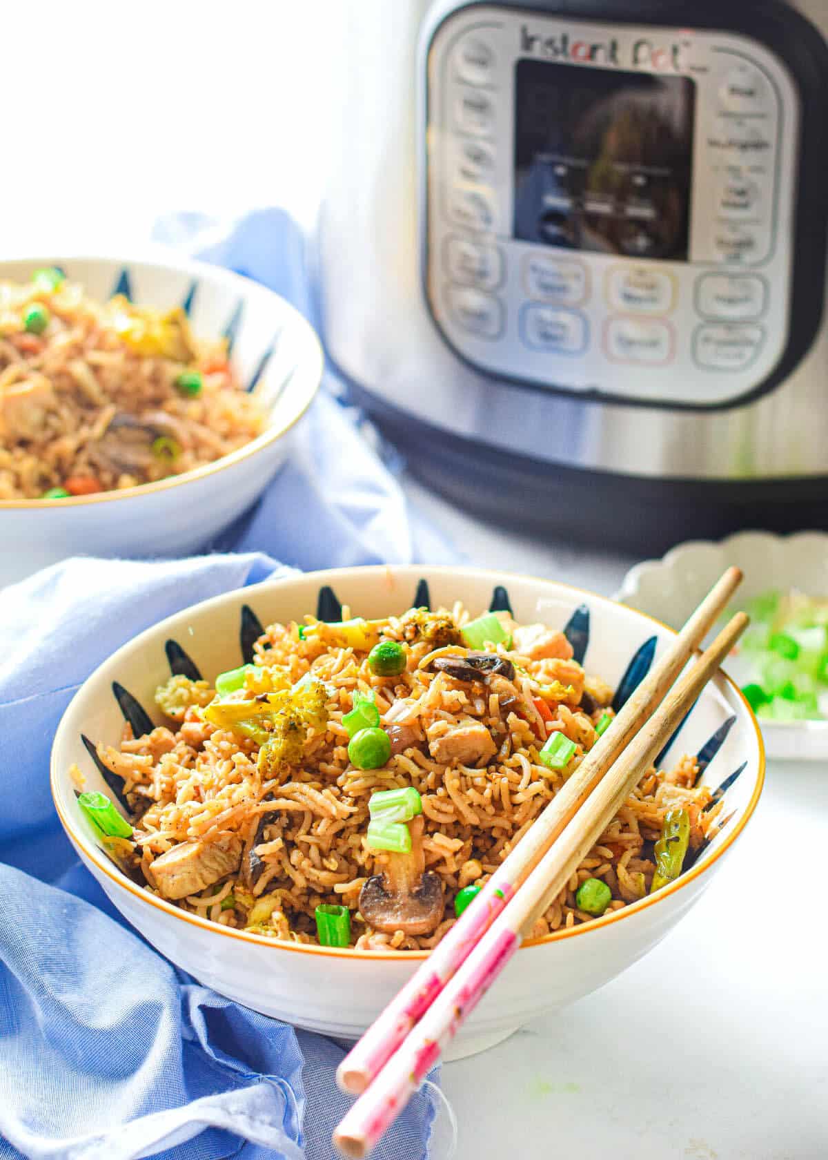 Instant Pot fried rice with chicken in a round white bowl with a pair of chopsticks on top, on top of a blue napkin, and an Instant Pot and another bowl of fried rice in the background.