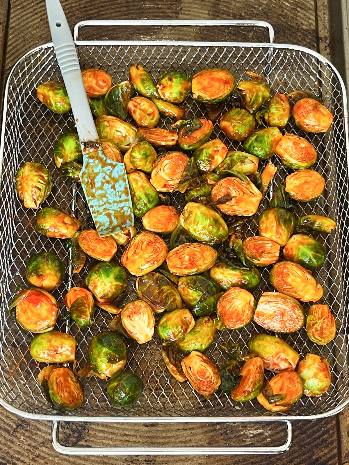 Brussels sprouts coated with a gochujang sauce sitting in an air fryer basket with a rubber spatula on top.