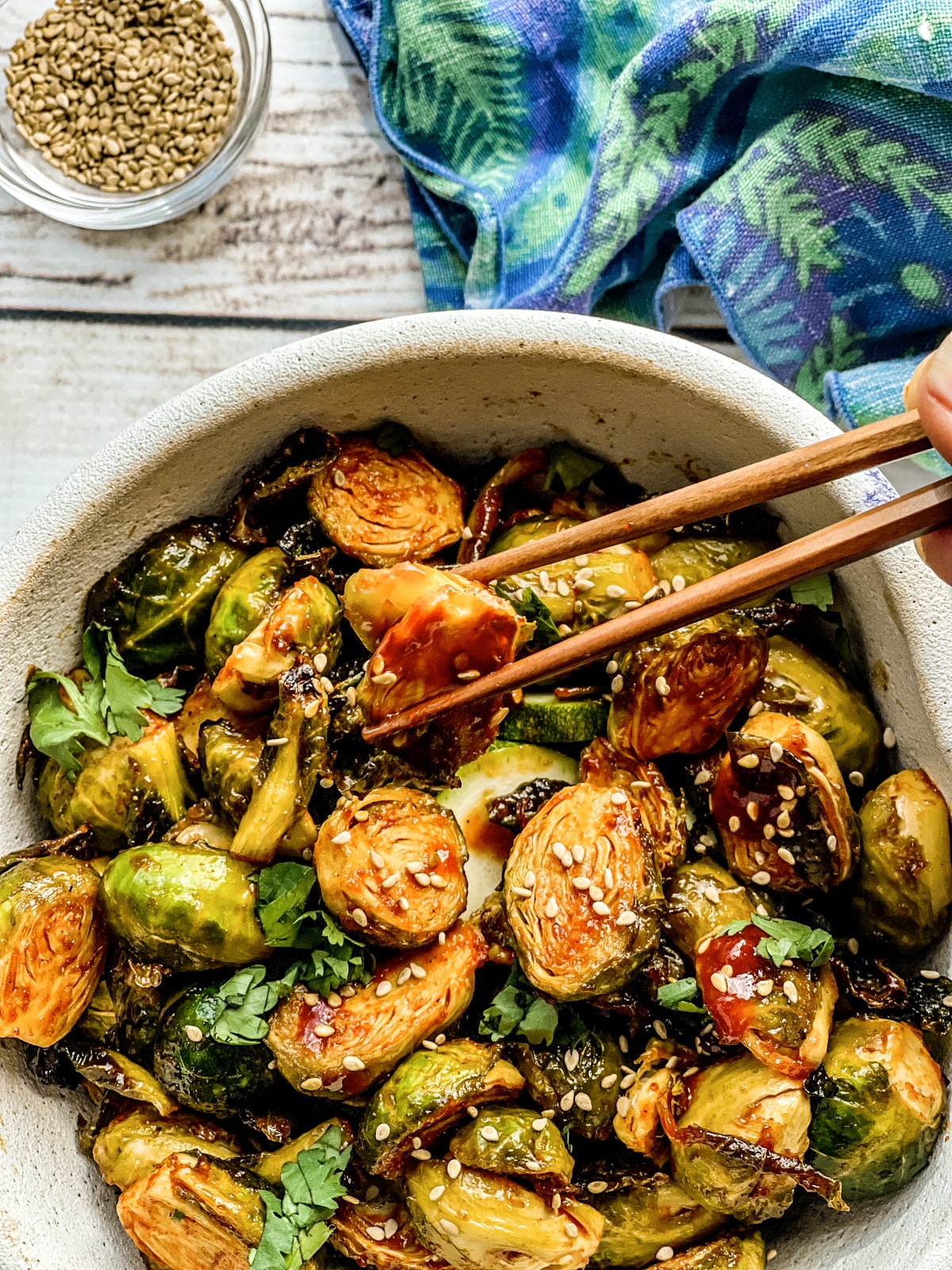 Chopsticks lifting up roasted brussels sprouts with gochujang sauce from a white bowl, with a blue napkin on the side.