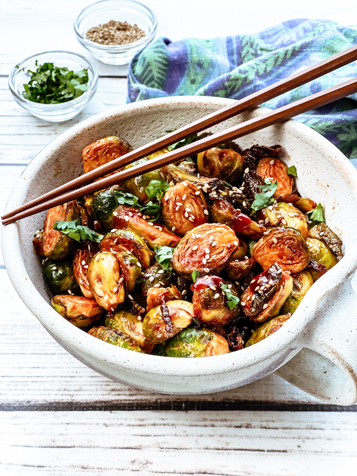 Air fryer brussels sprouts with gochujang in a white bowl topped with chopsticks with a blue napkin and garnishings in small bowls on the side on top of a white board.