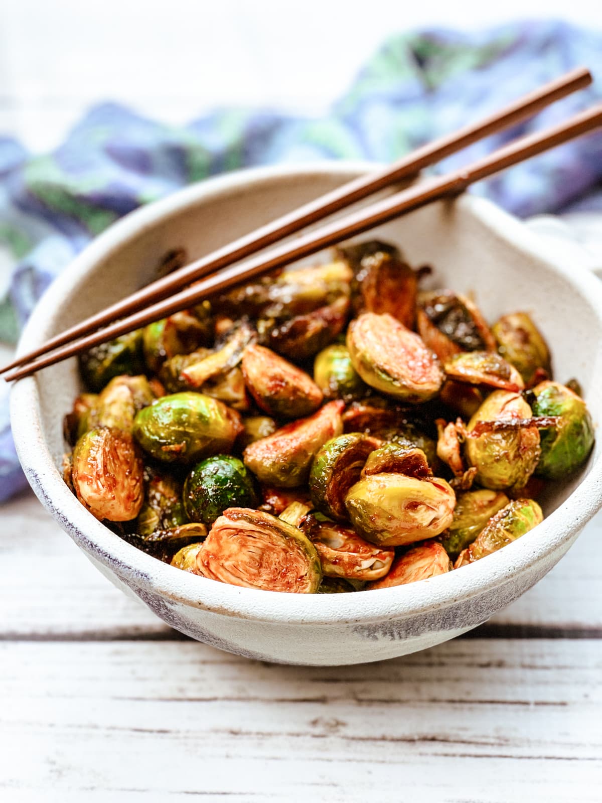 Air fryer brussels sprouts with gochujang in a white bowl topped with chopsticks with a blue napkin on the side on top of a white board.