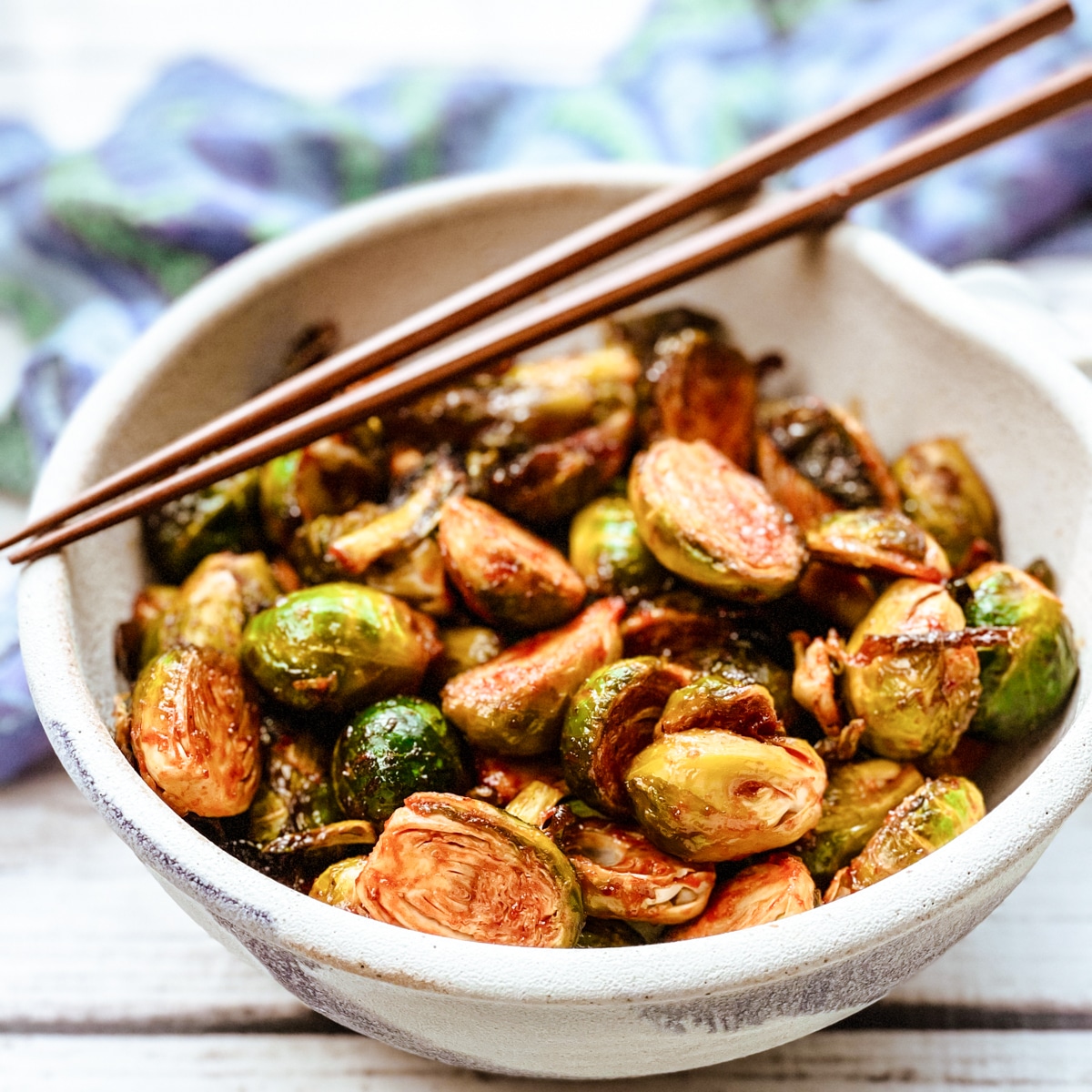 Air fryer brussels sprouts with gochujang in a white bowl topped with chopsticks with a blue napkin on the side.