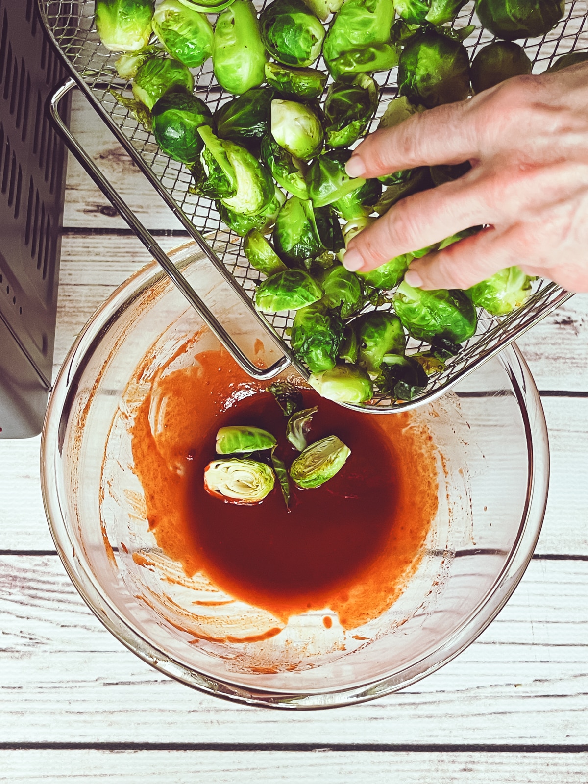 Raw, sliced brussels sprouts being poured into gochujang sauce a glass bowl on top of a white board.