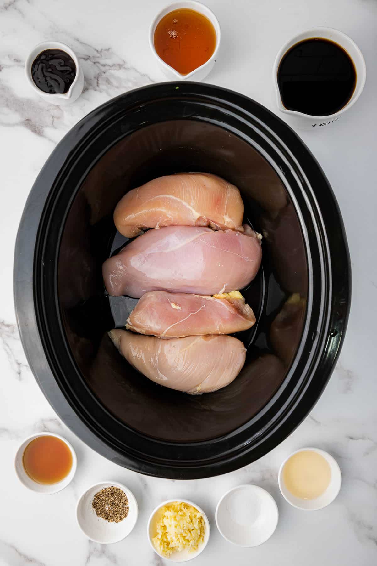 Four chicken breast fillets placed inside a slow cooker with ingredients for the honey garlic sauce in small white bowls around the slow cooker, on top of a marble surface.