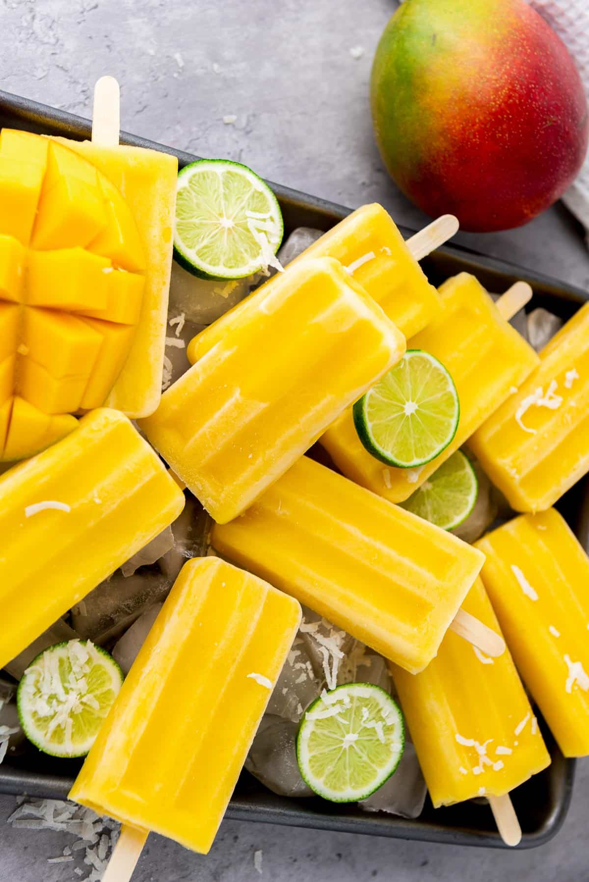 Mango coconut lime popsicles stacked on top of each other on a silver tray with lime slices on top and a whole mango in the background.