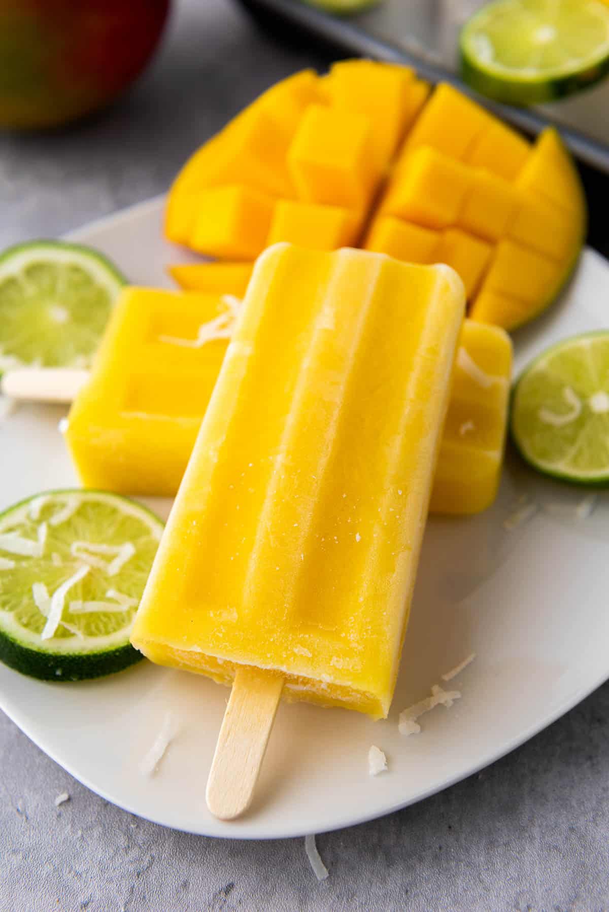 Mango coconut lime popsicles stacked on top of a white plate with lemon slices and a cut up mango, on top of a gray surface.