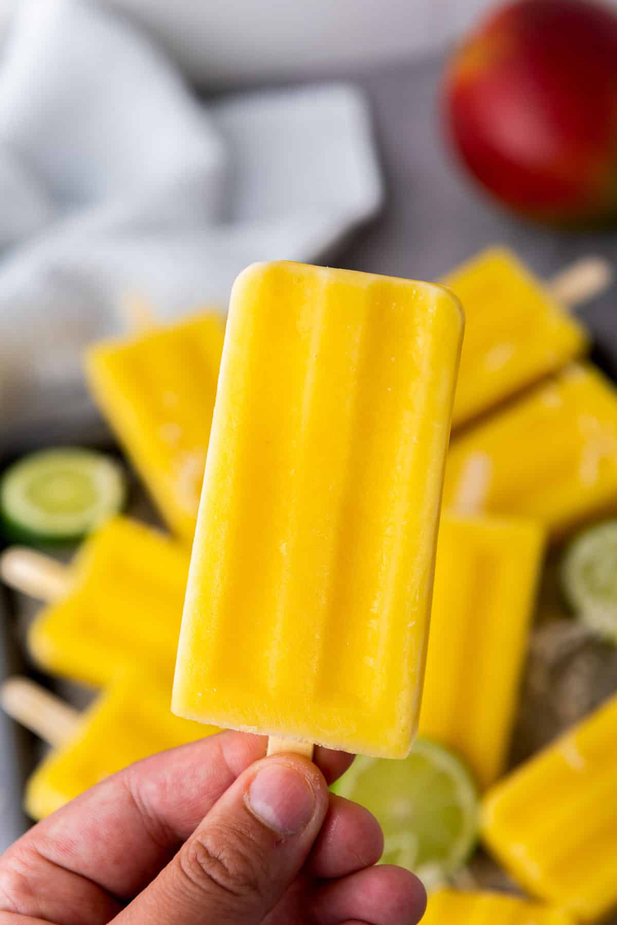 A hand holding up a mango coconut lime popsicle with more popsicles and lime slices underneath.