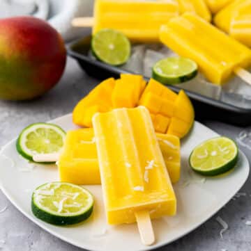 Mango coconut lime popsicles on a white plate with slices of lime on top and more popsicles in the background with a whole mango.