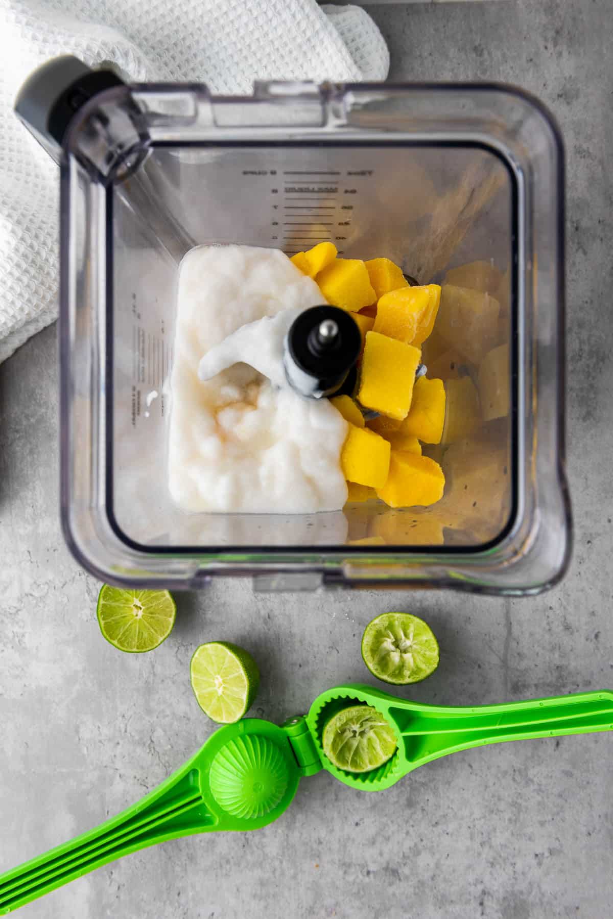 Chunks of mango and coconut yogurt in a blender with a lime squeezer and fresh limes on the side, on top of a gray surface with a white napkin on the side.