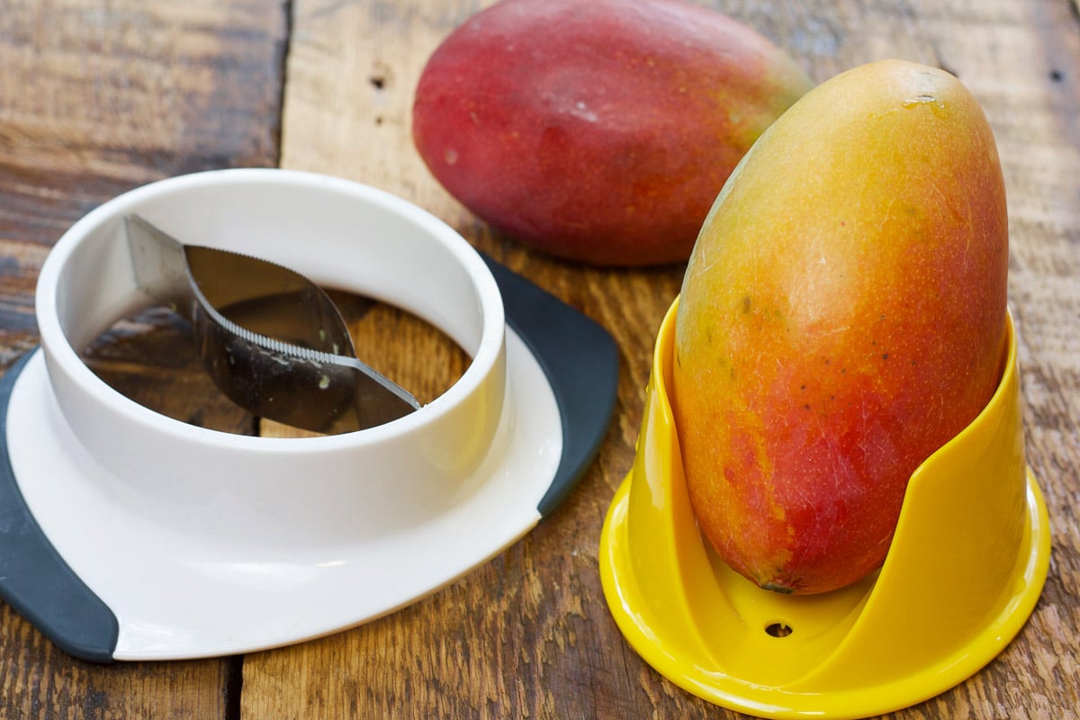 Whole ripe mangos on top of a wooden board with a mango cutter.