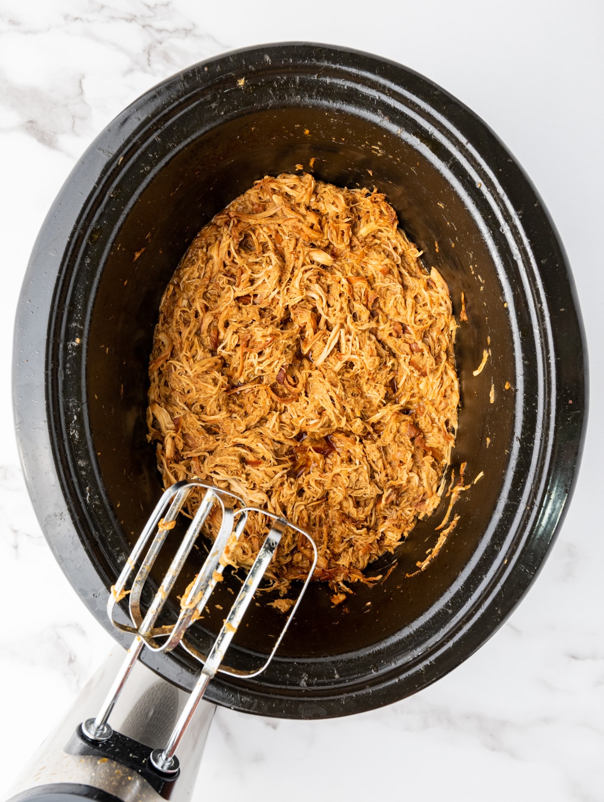 Shredded honey garlic chicken inside a slow cooker with a hand beater on the side, on top of a marble surface.