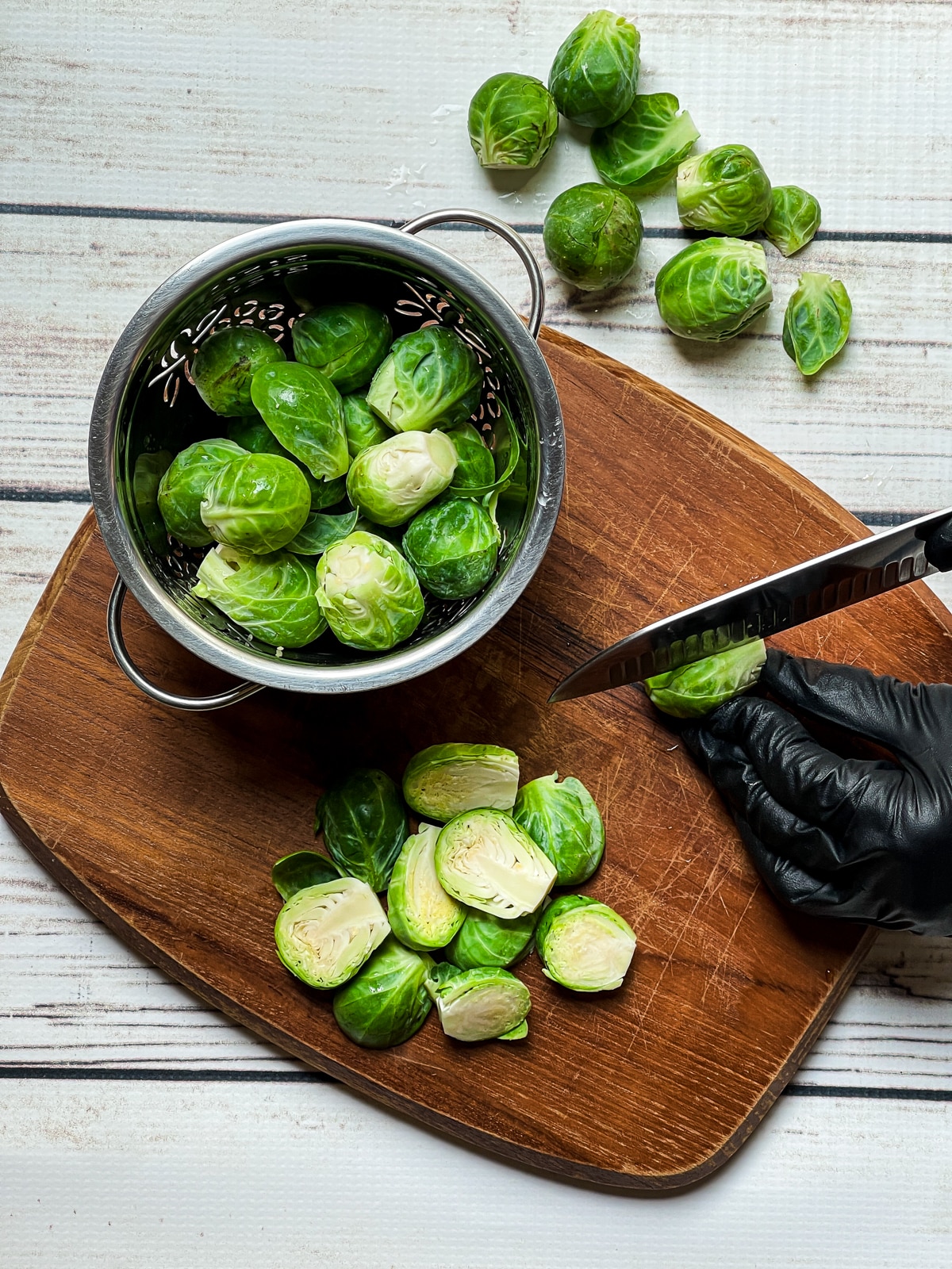 A hand slicing raw brussels sprouts in half on top of a wooden cutting board, with whole brussels sprouts on the side on top of a white board.
