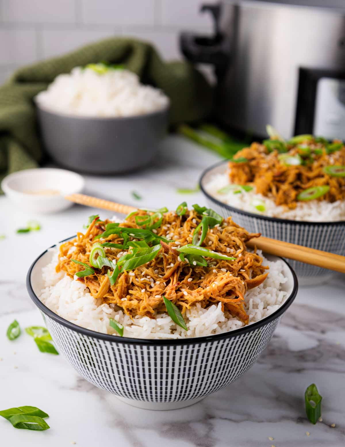 Slow cooker honey garlic chicken over white rice topped with sliced scallions placed in two white bowls with a pair of chopsticks on top and a small bowl of sesame seeds and a black bowl of white rice on the side, on a marble surface.