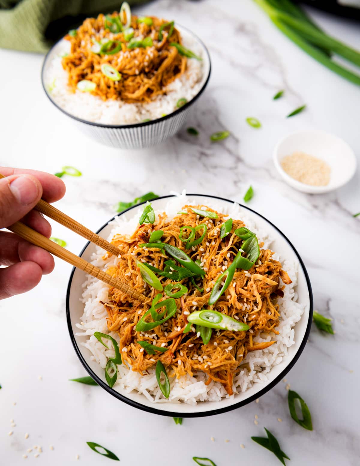 A hand using chopsticks for a bite of slow cooker honey garlic chicken over white rice topped with sliced scallions in a white bowl, and a small bowl of sesame seeds and a green napkin on the side, on top of a marble surface.